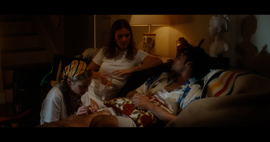 A group of three is gathered in a dimly lit room. A man is seated on a couch looking at a woman seated beside him on a lounge chair. Between them is a person crouched with their attention fixed on the thigh of the seated man, they are preparing to give him the injection shown in the previous picture